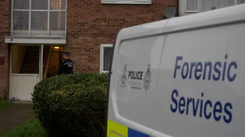 Jamie Niblock/BBC A white police forensic services van parked next to a building of flats. A policeman is standing next to the door. There is a green hedge between the van and the brick-built property. 