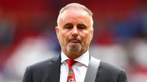 A bearded man wearing a red tie and suit inside a football stadium. He is playing a guitar