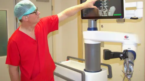 Dr Simon Tizzard is wearing a blue medical hat, orange scrubs and glasses pointing at a spine X-ray on the screen of the robot in a hospital room.
