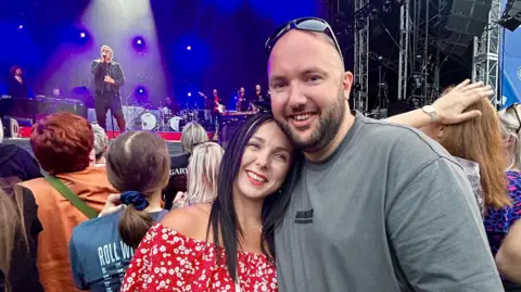 Cuffe and Taylor A woman puts her head on a man's shoulder as they both smile at an outdoor concert. Behind them on stage is Gary Barlow