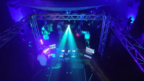 Sam Seven An aerial shot of a darkened nightclub, with the back of the DJ and their equipment in the foreground, and behind, people dancing under purple and green lights