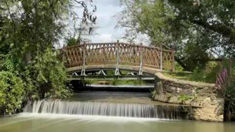 Esther J Water runs over a weir underneath a brown wooden bridge with a stone wall to the right and trees either side