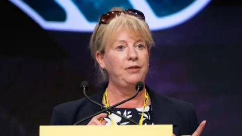 PA Media Shona Robison talks at a yellow podium with sunglasses propped on her head