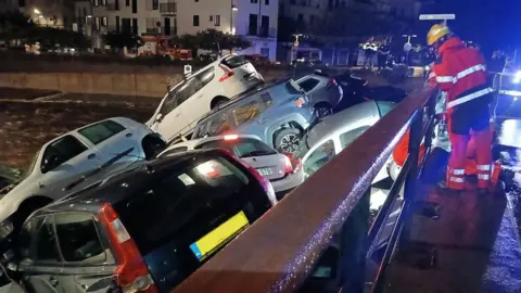 Catalonia fire service A fireman in a red suit with white reflective stripes and a yellow helmet looks down on a pile of cars, as water flows under them, at night time 