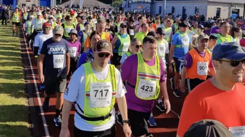 A large group of people in colourful walking gear wearing Parish Walk bibs leave the start line.