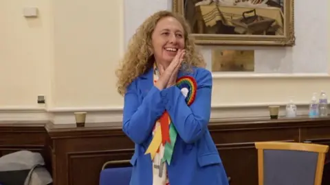 Inna Gardiner is stood up, smiling and holding her hands together in celebration, not looking at the camera. She has long curly hair, and is wearing beige trousers and a large Royal blue blazer with rainbow rosettes pinned on. There are chairs behind her. 