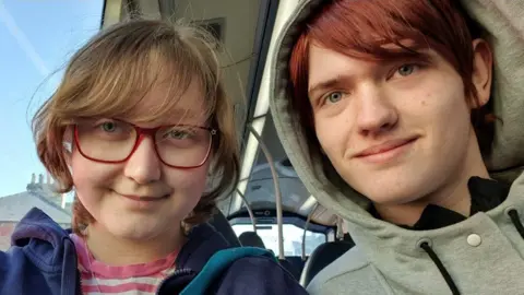 Supplied A young girl with fair hair and red-framed glasses on the left. She is wearing a blue hoodie and a pink and white striped T-shirt. On the right is a young man with red hair wearing a grey hooded top.