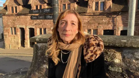 BBC A woman wearing a black coat and a brown scarf, stood in a churchyard in Ross-on-Wye. Behind her is a street sign.
