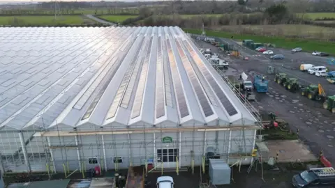 The image shows a large greenhouse with long rows of rooftop solar panels installed along its ridged roof. In front of the building is a yard area containing machinery and vehicles, including tractors and utility vehicles. Several cars are parked around the site. The surrounding landscape features open fields, hedgerows, and distant power lines.
