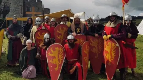 12 men and one woman in medieval costume. The men are in red soldier uniforms and the woman is wearing a pink skirt and green cloak. They are standing on a field