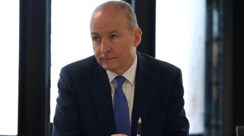 PA Media Taoiseach (Irish prime minister) Micheál Martin wears a blue suit, white shirt and blue tie. He has grey hair. He is sitting at a table.