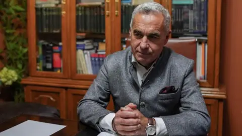 Reuters Georgescu, a man with grey hair and wearing a grey suit, sits at a desk, his hands clasped, looking pensive