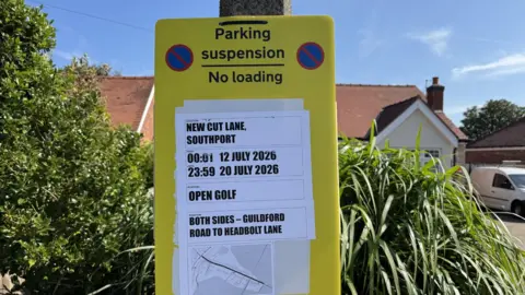 A parking suspension sign, it is yellow and has a big white area with writing on it about the closures. The sign is in front of a green bush.