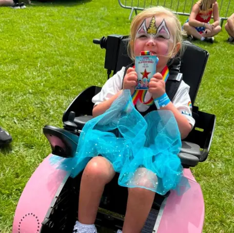 Warren Davies A young blonde girl with a unicorn design painted on her face. She is holding up a medal which is round her neck. She is wearing a white t shirt and blue tutu-style skirt and is sat in a black wheelchair with black straps holding her in. The wheelchair is on a grassy field, with other people sat on the field behind her.