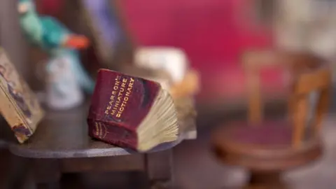 A table in a doll house with a burgundy and gold lettered book saying Pearson's miniature dictionary on it. The book sits on a grey table with a dark wooden chair with a burgundy cushion in the background which is blurred out.