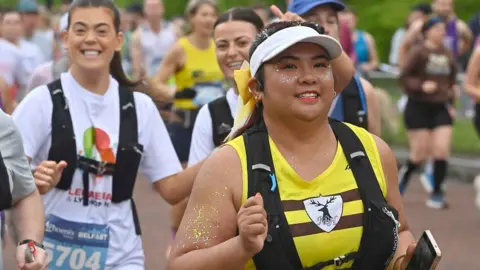 Runners at Belfast marathon - three ladies are smiling at camera. The main focus is a woman wearing a yellow vest and white visor, with gold body glitter