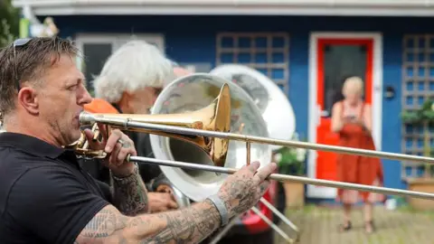 Penzance Council A musician with tattooed arms plays a trombone in the street as part of a lively parade, with other brass players and spectators gathered outside colourful buildings in the background.