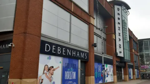 The exterior of the large Debenhams store in Sunderland. It has multiple storeys and still has adverts on the windows despite being closed since 2021. The store's name is above the windows, main entrance doors and up the middle of the building alongside a large glass frontage.