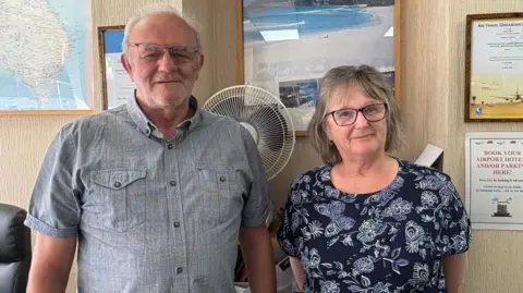 Kevin Shoesmith/BBC Travel agents Terry Robinson and Jo Pacey are standing in Louth Travel Centre. There is a picture of a beach, with turquoise sea, on the wall behind them.