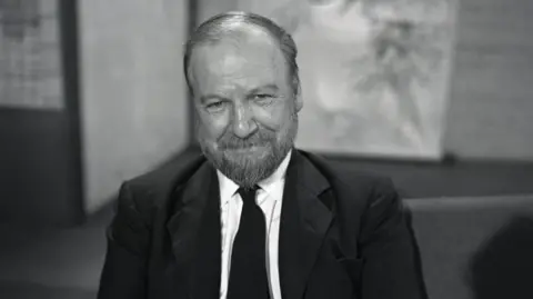 A black and white midshot image of William Golding smiling to camera and wearing a black suit jacket and black tie. The background is blurred