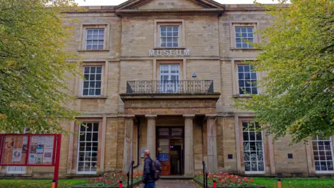 The façade of the museum includes the word 'Museum' on sand-coloured bricks. There is a balcony overlooking four columns above the entrance. A man is walking past the museum and there are trees on either side. 