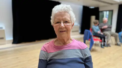 A white lady in her mid- eighties with grey hair and glasses, wearing a pink shirt with blue and white stripes.