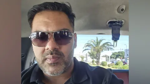 Afzaal Nasir Afzaal Nasir has black hair and a small beard. He is wearing sunglasses and a black T-shirt inside his taxi. He is in a sunny destination with palm trees in the distance outside the vehicle.