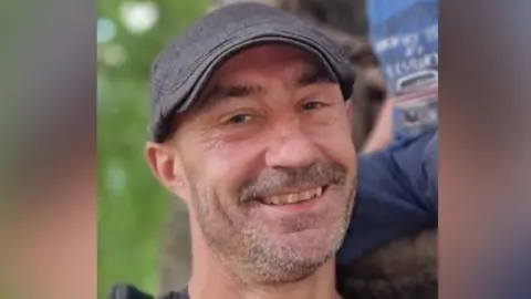 A man with salt-and-pepper stubble, wearing a dark-coloured jumper or t-shirt and a grey flat cap. He is smiling at the camera.