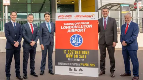 London Luton Airport Five men in suits looking towards the camera. Between them is a sign for Jet2.com, saying it is taking off from London Luton Airport in white lettering on an orange background. They are standing outside the airport, an entry to which can be glimpsed behind them