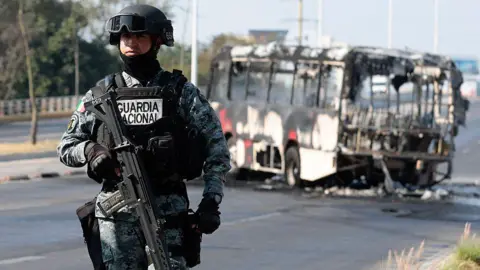 A National Guard member stands guard near a burnt bus set on fire by organised crime groups in response to an operation in Jalisco to arrest a high-priority security target, at one of the main avenues in Zapopan, state of Jalisco, Mexico, on February 22, 2026.