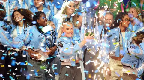 PA Media Manchester City's Alex Greenwood with the Women's Super League trophy with confetti floating around the players. The players are all smiling and laughing.