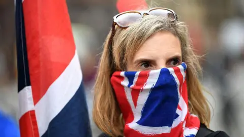 Getty Images A  woman with glasses perched on her head wears a union jack bandana that covers the lower part of her face. She has shoulder-length blonde/grey hair with  a fringe swept to the side and has a fixed stare. She is holding a union jack flag which is hanging down.