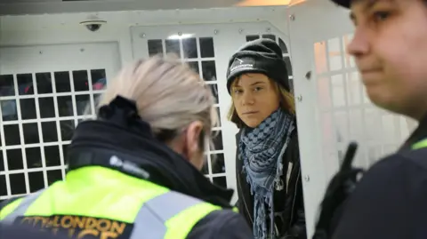PA Media Greta Thunberg inside a police vehicle, looking through a partition, with police officers visible in the foreground.
