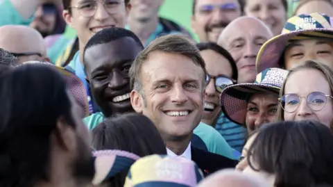 ANDRE PAIN/POOL/AFP Le président français Emmanuel Macron pose avec des bénévoles lors de sa visite à la compétition de tir à l'arc lors des Jeux olympiques de Paris 2024 sur l'Esplanade des Invalides à Paris le 2 août 2024.