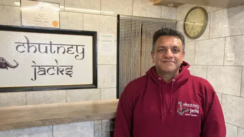 Amy Holmes/BBC A man in a maroon hoodie stands inside a tiled restaurant reception area beside a sign reading “chutney jacks.”
