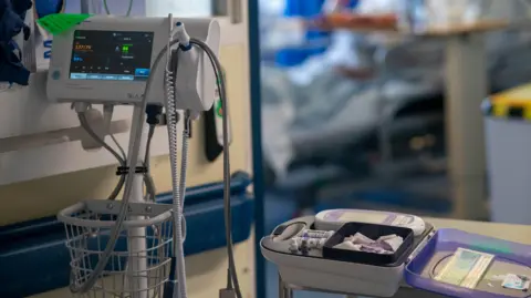 PA Media Hospital equipment with a monitoring screen and a bed with what appears to be needles and further monitoring equipment on it. A hospital room, including a bed, can be seen in the blurred background. 