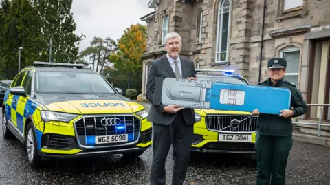 PSNI John O'Dowd, wearing a black suit and grey shirt, holds a large drug testing kit next to a uniformed policewoman in front of two police cars.