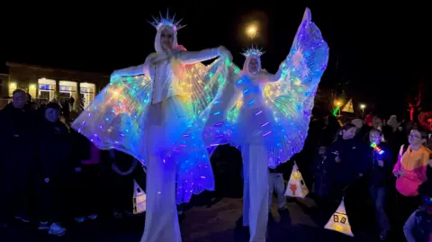 BBC Stilt walkers wearing bright costumes lit up in blue walk among crowds.