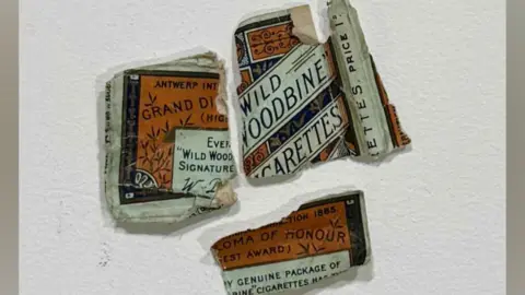 North Northamptonshire Council Ripped up cigarette packaging, that is three pieces, on a piece of white paper. It is white, brown and orange in colour. 