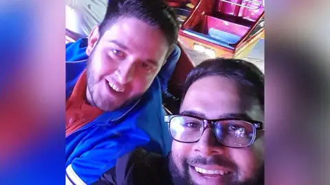 Selfie of the late Simon Walker (left) who has dark brown hair and stubble and wearing a blue jacket and orange shirt with Maj Mahmood (right) with black hair and beard and wearing dark brown glasses. They are sitting on what appears to be a ride and both are smiling. 