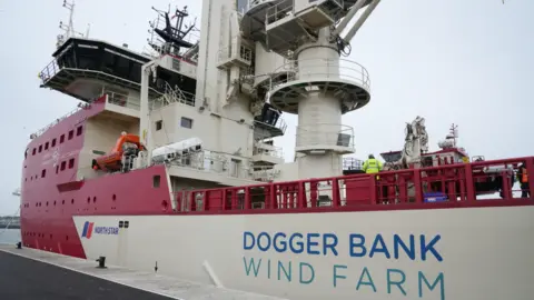 John Fairhall/BBC A large ship with the words Dogger Bank Wind Farm is docked at a port. It is red and white