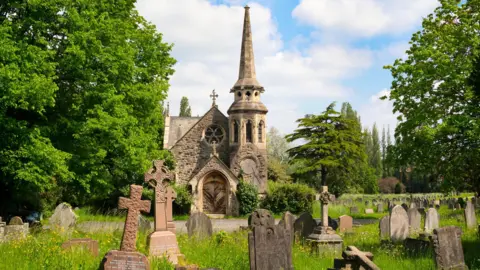 Getty Images An old chapel sits among grave markers and mature trees. The sun is shining and there are clouds in the sky. 