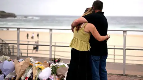 Mourners embrace near tributes piled together in memory of the victims of a shooting at Bondi Beach. A woman wearing a yellow top and black skirt hugs a man wearing a black polo and blue jeans as they look at a pile of flowers. In the background Bondi beach can be seen with people walking on the sand and sea in the distance