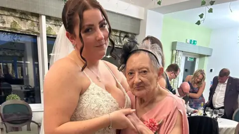 Lauren Adamson is pictured dancing with her grandmother Margaret Harris during her blessing ceremony celebrations. Lauren is wearing a white wedding dress with floral details on it. Her brown hair has been tied back with two pieces styled around her face. She is holding Margaret close to her and holding her hands. Margaret is wearing a pink dress with a black fascinator. They are both smiling at the camera.