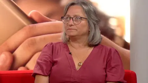 A woman in a maroon dress sits on a red sofa. She wears black glasses and has shoulder-length grey hair.