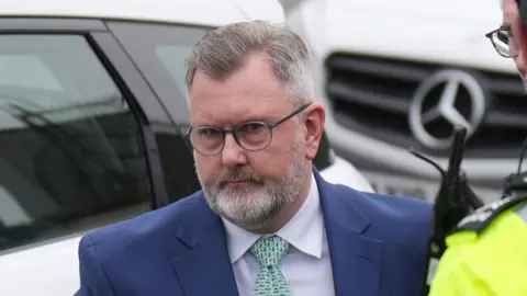 PA Media A man with a white beard, darker moustache and round glasses wearing a dark blue suit, white shirt and green tie