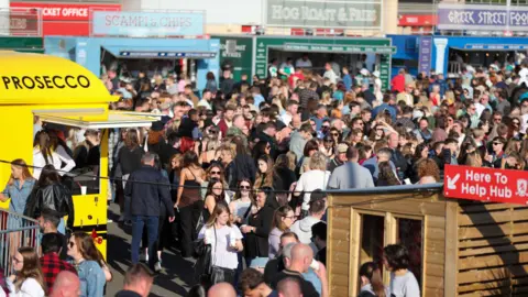 Crowds of people at a food market in Middlesbrough. Food vans surround the square selling various food. A yellow van to the left advertises prosecco. Three vans to the back of the crowd are selling scampi and chips, hog roasts and Greek street food.