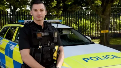 Avon and Somerset Police Sgt Luke O'Connell in police uniform standing in front of his marked police car