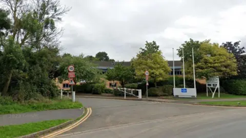 The entrance to office block Ashford House in Walsgrave Triangle Business Park. The picture is taken from the road opposite the entrance, with several trees lining the pavements and a building largely obscured by the foliage in the background.