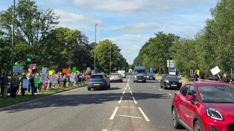 A busy road with protesters with placards and signs lining the grass verge